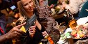 Meat on a large skewer sliced infront of people in a busy restaurant
