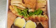 A boxed assortment of afternoon tea items, including sliced sandwiches with ham and cheese, two sausage rolls, croissants with salad and cheese, two s