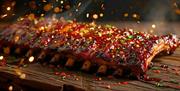 A rack of glazed, barbecued ribs sits on a wooden surface, topped with chopped herbs and chili flakes, with glowing embers and sparks in the air, evok