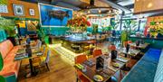 Interior of The Brazilian. A brightly lit, modern restaurant interior with colorful seating, set dining tables, and vibrant floral arrangements. The s