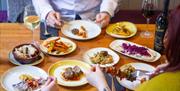 Wooden table with various Tapas on plates hands holding knives and forks.