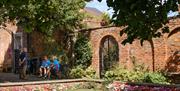 A brick-walled garden with pink and white flowers, two gated archways and a group of people sat on a nearby bench.