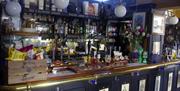 A well-stocked pub bar with beer taps, bottles of liquor, a wooden crate of crisps, a pint of beer, a bouquet of flowers, and various decorative eleme