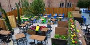 Outdoor seating area with several tables and chairs, some greenery, and a person tending to a planter with flowers. Brick walls and wooden fencing enc