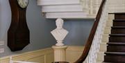 An ornate  white staircase with darkwood flooring and banister, alongside an antique wallclock, a marble bust of William Wilberforce and a black and w