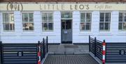 Exterior of "Little Leo's Café Bar," a white-painted building with large windows and a light beige sign above the entrance. In front of the café, ther