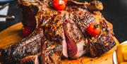 A large, grilled tomahawk steak on a wooden cutting board, garnished with roasted cherry tomatoes and red peppercorns, with a side dish of roasted pot