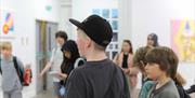 Several children and teenagers are gathered in a brightly lit art gallery, looking at the artwork displayed on the white walls.