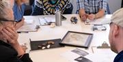 A group of six people sit around a white table engaged in discussion, with various items on the table including rocks, a framed map, papers, a water b