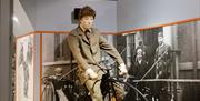 A mannequin of an early 1900s man sits on a penny farthing bicycle with photographic displays of victorian imagery behind him.