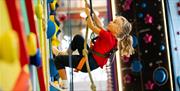 A small child in a harness smiles as they pull a rope to help themselves climb a multicoloured climbing wall.