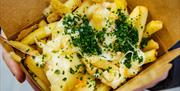 Fries in a cardboard takeaway carton, covered in cheese topped with green herbs.