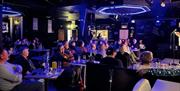 Crowd sat at tables inside a comedy club laughing while watching a performance on stage.