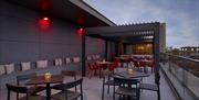 Exterior of The Lexington Bar. A modern rooftop terrace with black tables and chairs, red accent chairs, and a long bench with white cushions along on