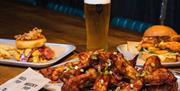 Chicken wings on a white plate with burger and a pint of beer on a wooden table.