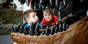 2 young boys on their knees look cautiously up into the open mouth of a lifesize dinosaur mouth.