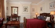 A cosy bedroom with a double bed covered in a deep red comforter, several decorative pillows, two matching wooden nightstands with lamps, a wooden cof