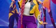 A person dressed in a colorful outfit with a long pink braided wig poses energetically in front of a backdrop featuring three animated characters in s