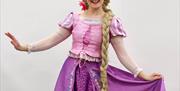 A person dressed as Rapunzel from the Disney movie "Tangled" poses with a big smile, wearing a long blonde braided wig and a purple and pink dress. A
