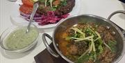 A bowl of spicy meat curry garnished with sliced green chilies, ginger, and cilantro is placed on a wooden board. Next to it is a dish of kebabs or pa