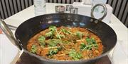 A black metal pan filled with a rich, red-orange curry garnished with fresh cilantro and thin slices of ginger sits on a wooden board; in the backgrou