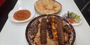 A table set with a plate of Afghan mixed grill including rice, kebabs, grilled meat, and carrots, accompanied by a bowl of lentil soup, a round flatbr