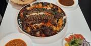 A white table set with a large platter of mixed grilled meats and rice in the centre, surrounded by plates of naan bread, chickpea curry, dark lentil