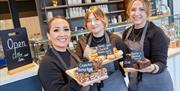 Three smiling café staff members wearing gray aprons stand behind the counter, each holding a tray with baked goods labeled "Crunchie Rocky Road," "Ch