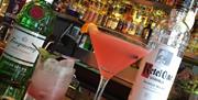 Two mixed drinks are displayed on a bar counter, one in a short glass and the other in a martini glass with a pink cocktail and an orange garnish, fla