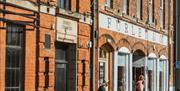 People sit at outdoor tables enjoying drinks and conversation in front of a red brick building with sign reading and "Furley & Co." on a sunny day.