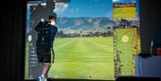 A young man in shorts swings a golf club on a small turf, with a projection of a golf course in front of him.
