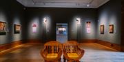 2 wooden chairs sat in the middle of an art gallery, with several pictures lining the walls.