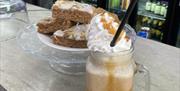A glass mug filled with a frothy liquid, topped in whipped cream and caramel sauce with a straw in, sat next to a plate of cakes on a glass display un
