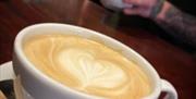 A close-up of a cup of latte with heart-shaped latte art on top, served in a white cup labeled "SLEEPERS CAFE BAR" on a saucer with a brown sugar pack