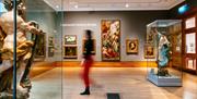 A blurred woman wearing red trousers walks through an exhibit with paintings on the walls and 2 statues of men in the middle of the room.