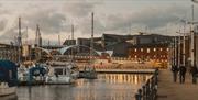Hull marina at dusk with moored boats and tall masts reflected in calm water, a pedestrian walkway along the quay, and modern city buildings illuminat