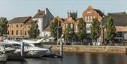 Motorboats moored along Hull marina in front of red-brick waterfront buildings and trees, with calm water in the foreground and historic city architec