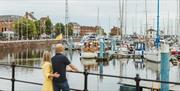 Two people stand by a railing overlooking Hull marina filled with moored boats and yachts, with calm water, masts, and red-brick buildings lining the