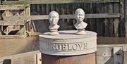 Two white sculpted busts displayed on a circular structure engraved with the word “TRUELOVE,” set against a backdrop of weathered wooden dock walls an