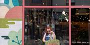 A person wearing sunglasses and denim overalls stands in front of a shop called "Plant & Paint," holding a green tote bag with the words "BOTANIC 'ULL