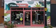 Colourful storefront for a shop called "Plant & Paint" with a pink door and large windows displaying potted plants, art prints, and t-shirts. The exte