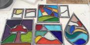 Several colourful stained glass panels of various shapes and designs are laid out on a white work surface at Studio Eleven. The panels feature abstrac