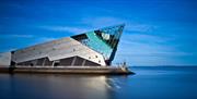 An angular, modern building with metallic and glass surfaces sits on the edge of calm water under a clear blue sky. The Deep features sharp, geometric